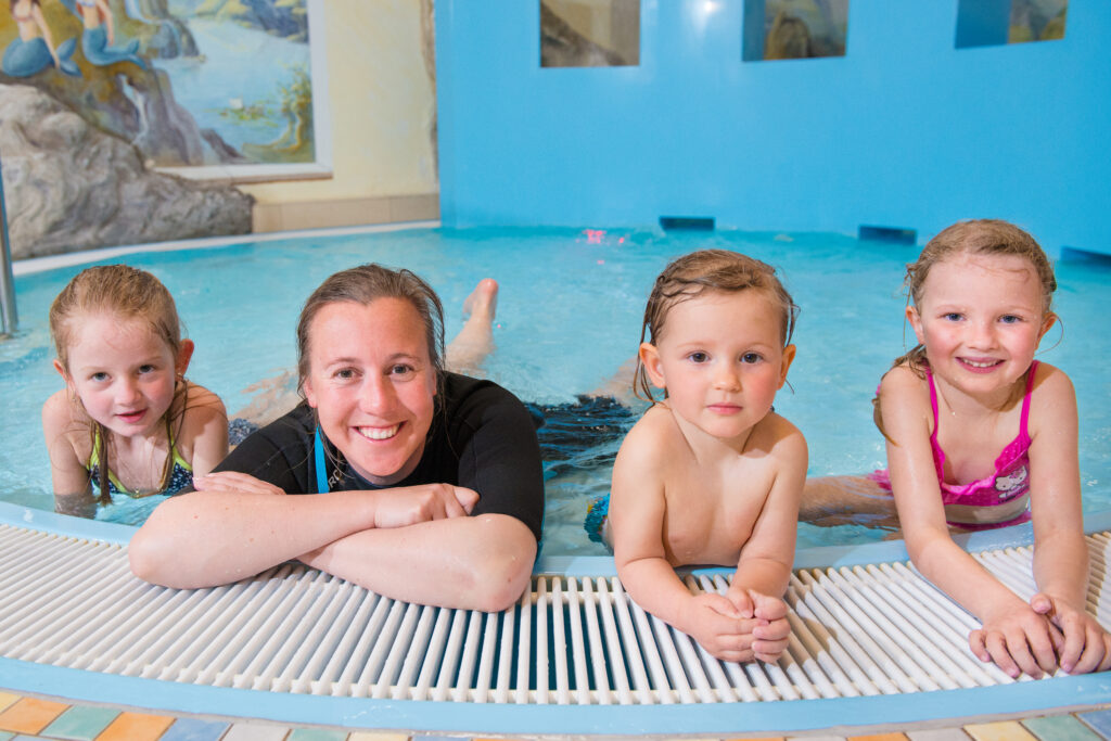 Unsere Schwimmlehrerin und drei Kinder liegen am Rand eines Schwimmbeckens in einem Innenbereich. Familienhotel Zauchenseehof