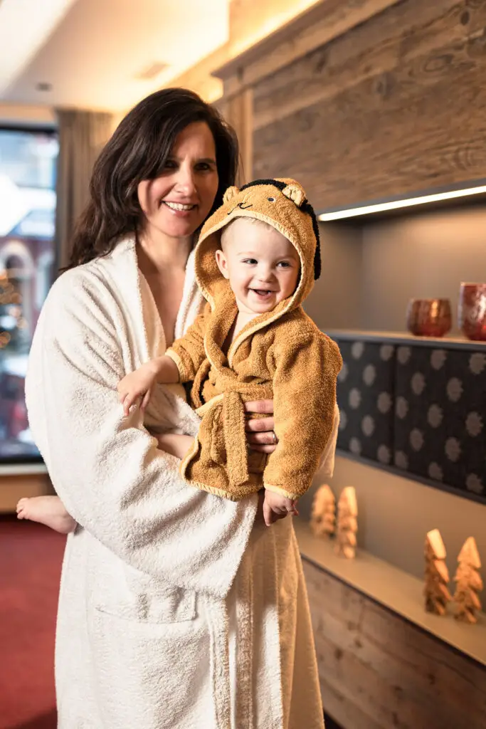 Eine Frau trägt ein Baby in einem Bademantel, beide lächeln in die Kamera. Familienhotel Zauchenseehof