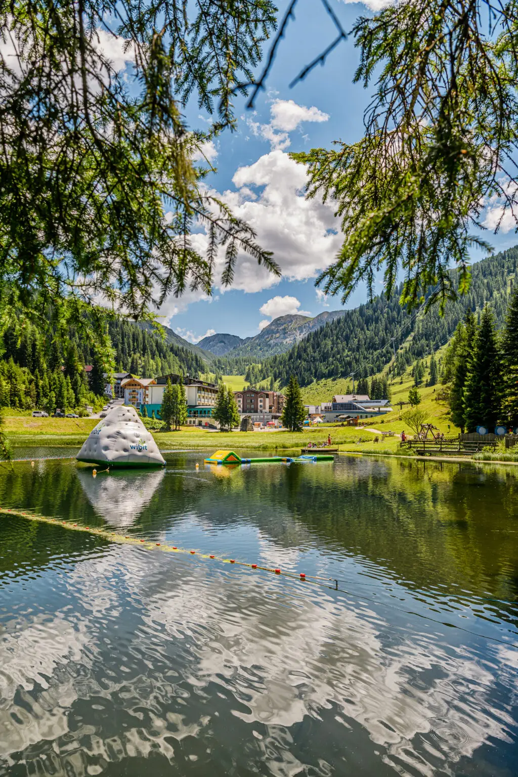 Im Zauchenseehof in Zauchensee spiegelt sich ein Badesee vor gr&uuml;nen Bergh&auml;ngen, w&auml;hrend ein Wibit-Element im Wasser liegt. Familienhotel Zauchenseehof Salzburger Land